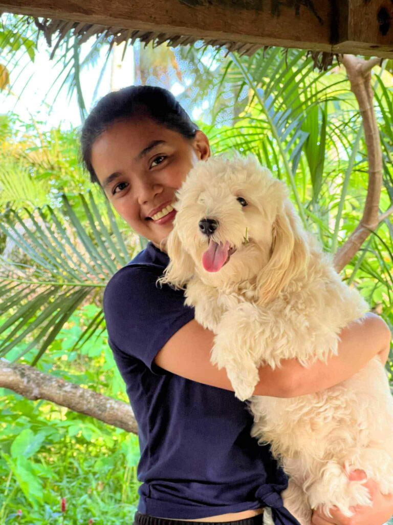 Smiling female seafarer AB Cherry Opena holding a fluffy white dog in a tropical outdoor setting with lush green palm trees in the background.
