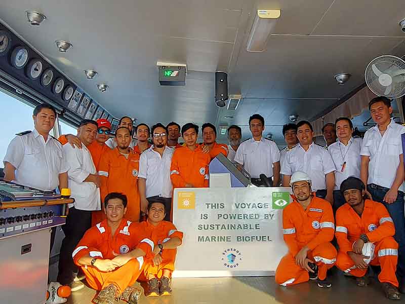 Crew members wearing orange coveralls and white uniforms pose on a ship&rsquo;s bridge around a sign reading &ldquo;This Voyage Is Powered By Sustainable Marine Biofuel,&rdquo; promoting renewable energy use at sea