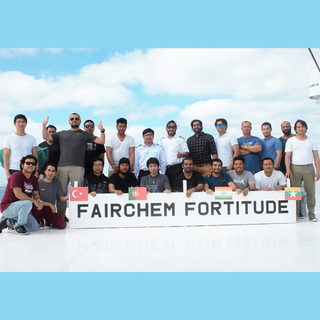 青空の下、「FAIRCHEM FORTITUDE」の甲板でポーズをとる多様な船員たちのグループ。